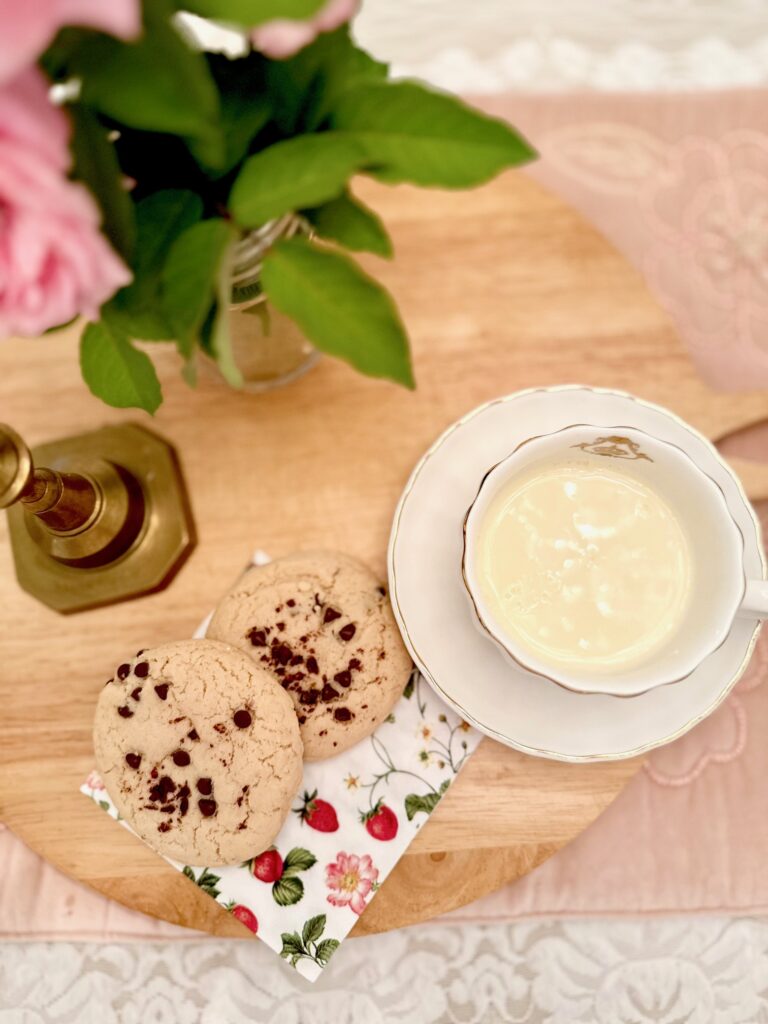 a lovely vignette two chocolate chip cookies gently placed on top of a strawberry napkin. The softest creamy milk in a white ceramic cottage core cup